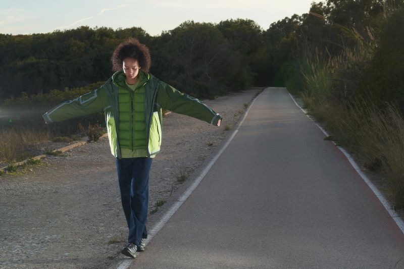 Person in green jacket balances on the edge of an asphalt path, with trees in the background, lit by sunlight.
