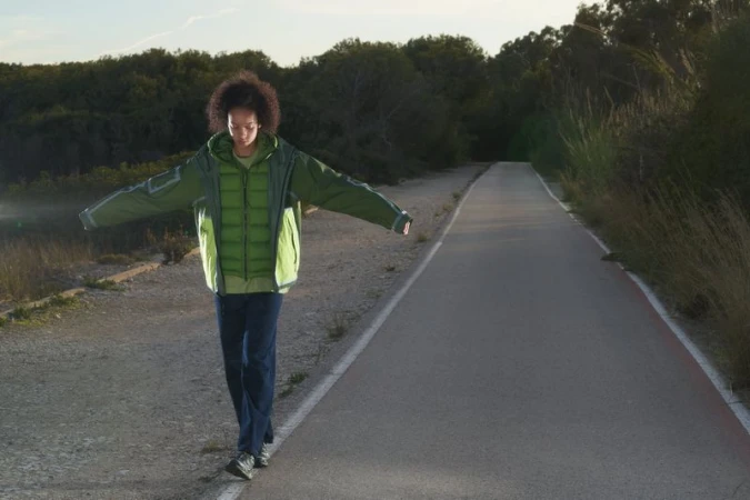 Person in green jacket balances on the edge of an asphalt path, with trees in the background, lit by sunlight.