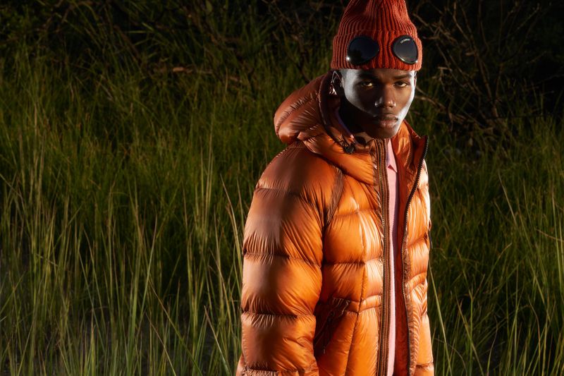 An adult male in a puffy orange hooded jacket, orange knit beanie, and goggle-style glasses poses in a natural setting at dusk.