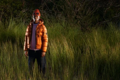 Young man wearing an orange puffer jacket and beanie standing in tall grass at night