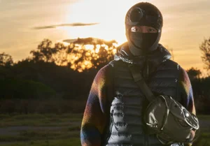 Person wearing a black balaclava with built-in goggles, a padded puffer vest, and a crossbody bag against a sunset sky.