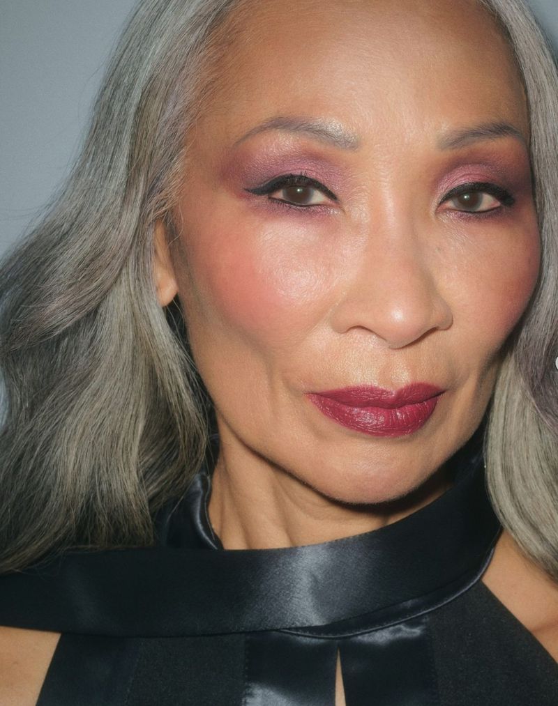 Close-up studio shot of an Asian adult female with gray hair, wearing pink eyeshadow, dark lipstick, and a black satin top.