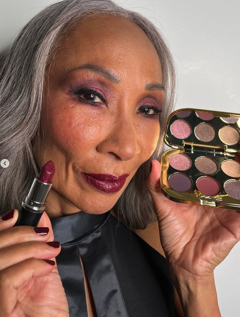 Close-up of an older woman with makeup holding lipstick and eyeshadow palette, showcasing a beauty product against a simple backdrop.