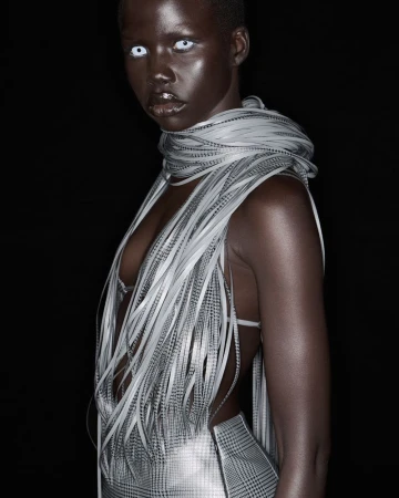Dark-skinned female model with white contact lenses wears a silver bra top and skirt with a feathered scarf against a black background.