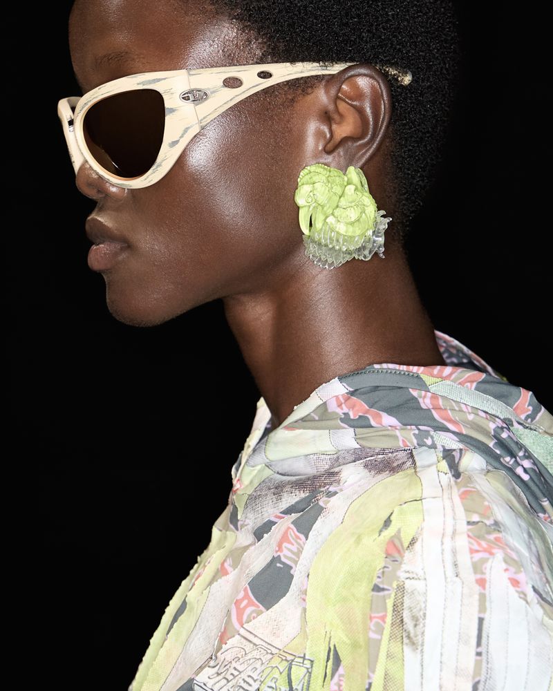 Close-up profile of a black woman wearing light-colored patterned sunglasses and a bright green floral earring for Diesel SS25.