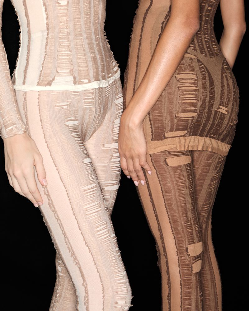 Two women wearing matching distressed beige and brown long sleeve tops and leggings posing against a black background in a studio shot.