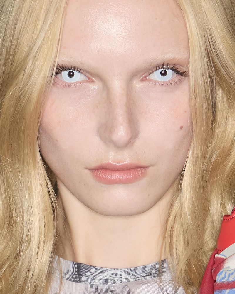 Close-up portrait of a blonde woman with pale skin and striking white contact lenses, highlighting her face and the color contrast.