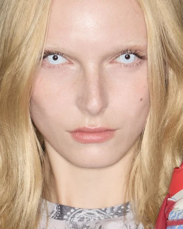 Close-up portrait of a blonde woman with pale skin and striking white contact lenses, highlighting her face and the color contrast.