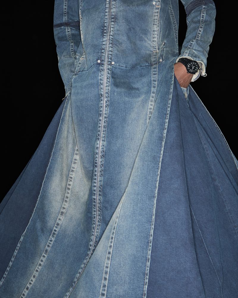 Fashion-forward denim trench coat with unique paneled construction and a single adult with a wristwatch on a black backdrop. 