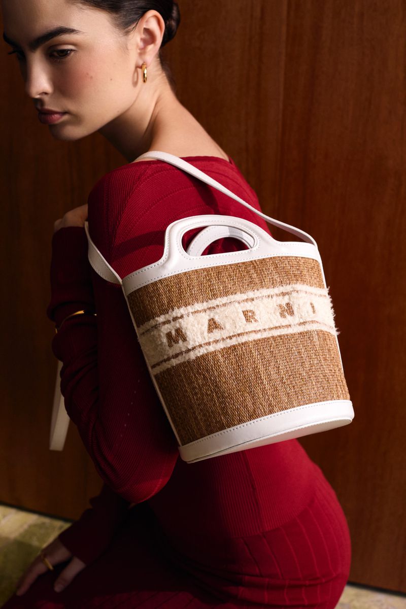 Model wearing a red top carrying a textured beige handbag with white trim against a wood paneled wall