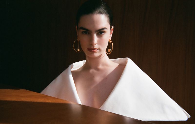 Portrait of a brunette woman wearing a white dress and gold earrings, posing indoors against a dark wooden background.