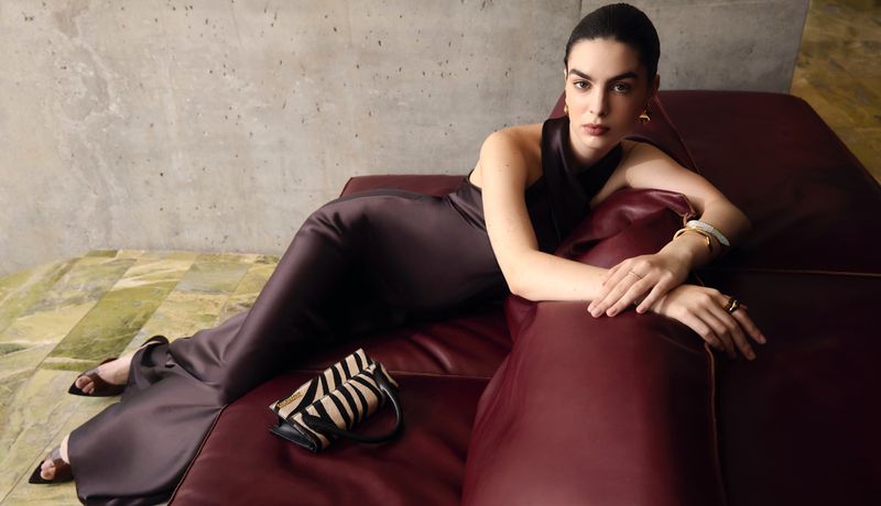 Adult woman in a satin dress reclines on a leather couch indoors with a zebra-print purse, accessorized with jewelry and black high-heeled shoes.