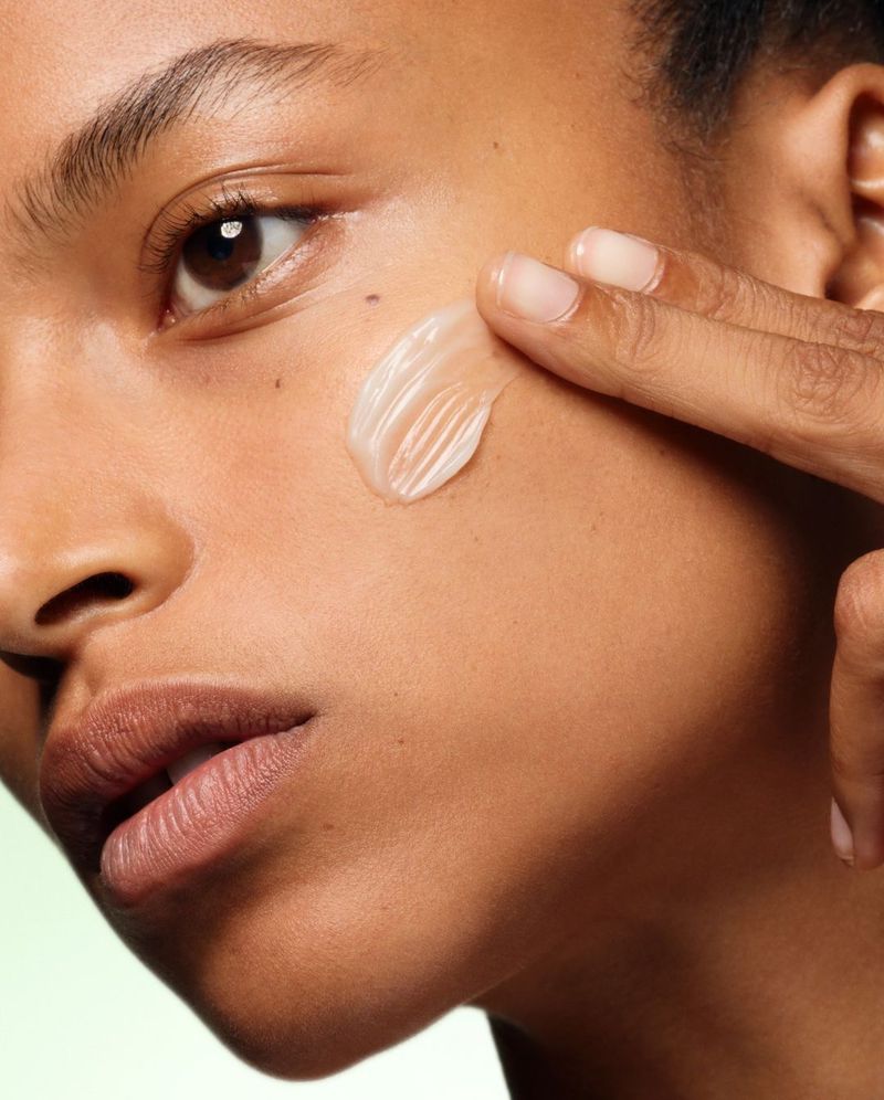 Close-up of an adult woman applying cosmetic cream to her cheek with her finger, highlighting skincare and beauty themes on a light green background.