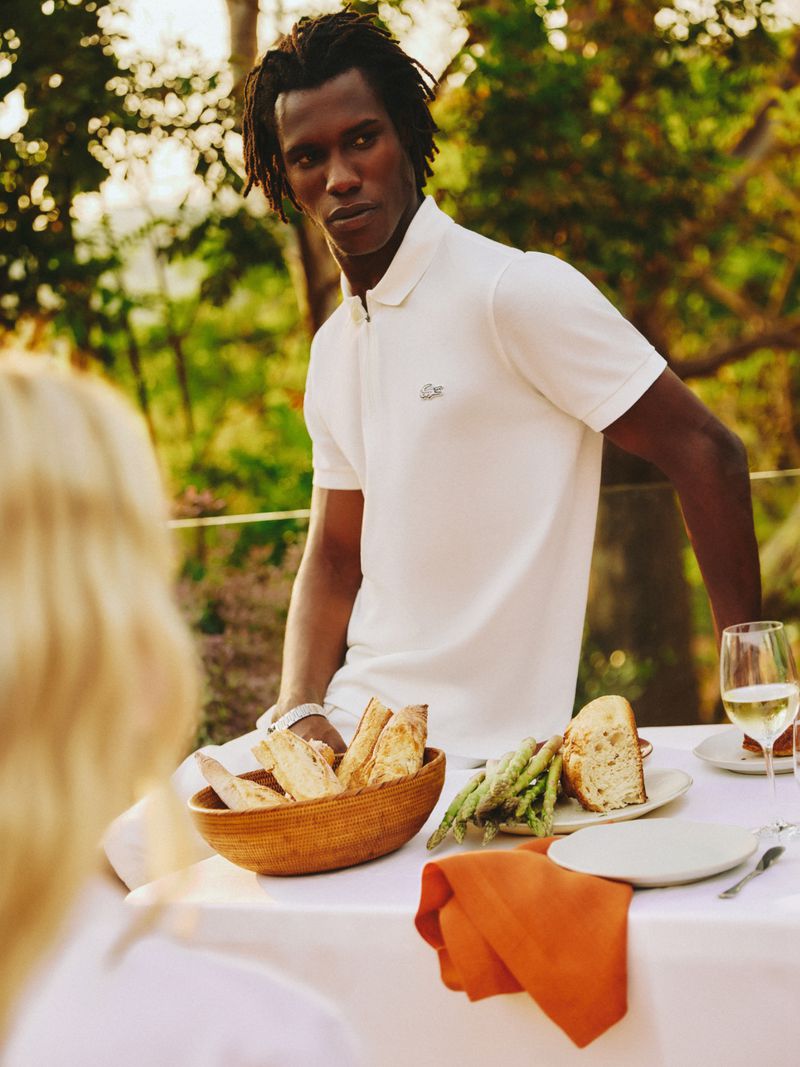 A man poses at an outdoor table with bread, asparagus, and wine. He's wearing a white polo shirt, and soft golden light fills the scene.