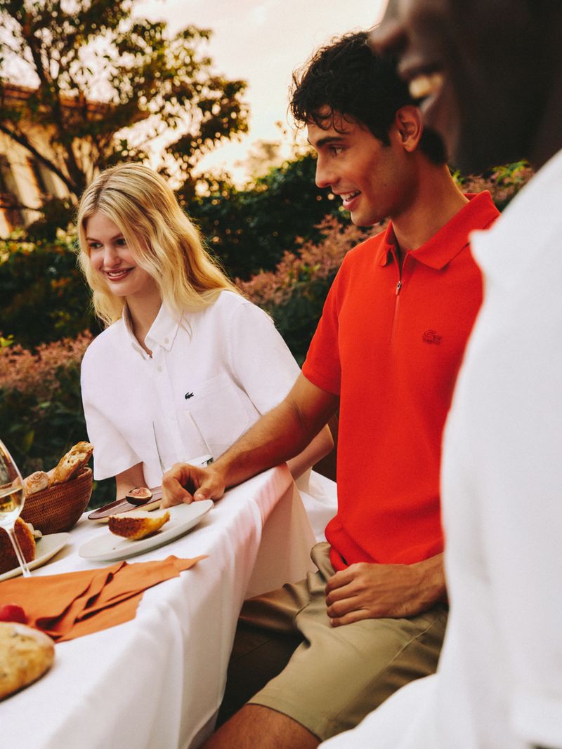 Three people sit at an outdoor table, enjoying food and drinks. A fair-haired woman smiles, next to a dark-haired man in a red polo.