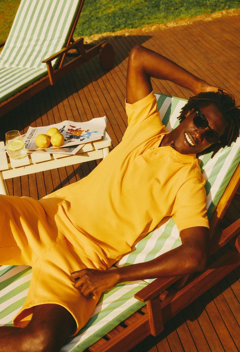 A smiling man relaxes on a striped deck chair wearing an orange shirt and shorts, sunglasses, with a drink and lemons nearby.