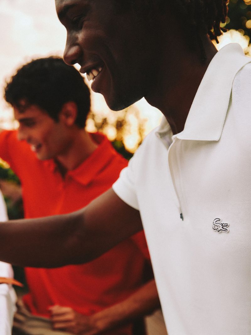 Close-up of two adult men smiling, one African-American wearing a white polo shirt with logo, the other a blurred Caucasian man in a red shirt.