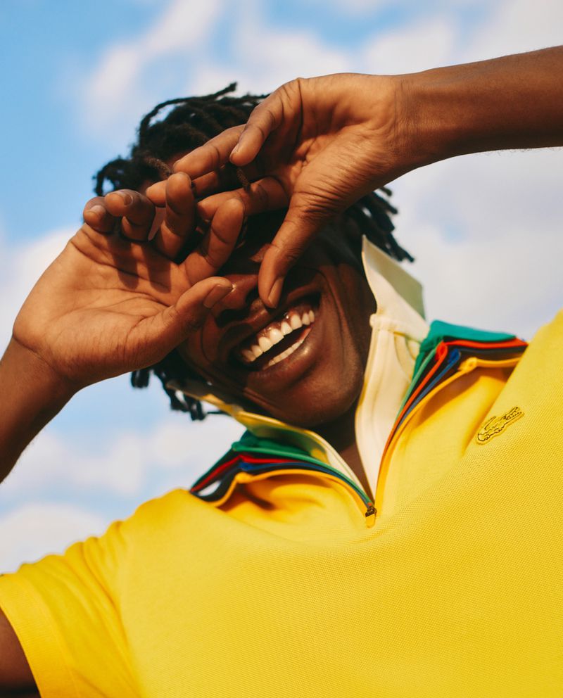 Smiling Black man shielding eyes in bright yellow Lacoste shirt against blue sky. Close-up headshot in direct sunlight showing joy.