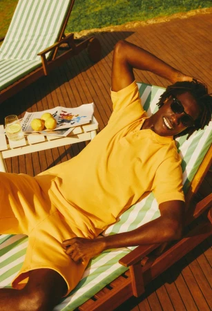 Black man wearing a yellow polo shirt and shorts relaxing on a striped lounge chair outdoors in sunlight for Lacoste Brazil Summer Pack 2024