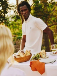 Black man wearing a white polo shirt standing by a dining table outdoors for Lacoste