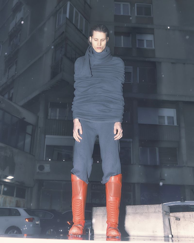 Fashion shot of a person wearing a layered grey top, matching pants, and red knee-high boots against an urban backdrop at night.