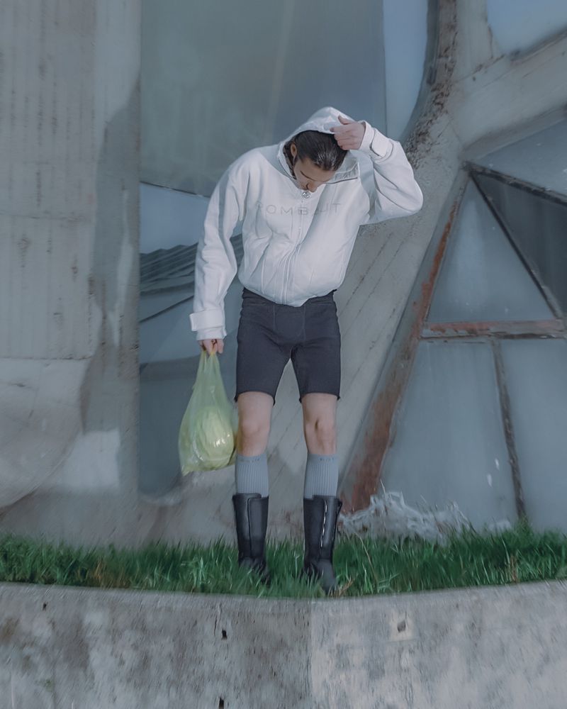 A person wearing a white hooded jacket, shorts, socks, and boots stands on a concrete structure holding a bag with a cabbage.