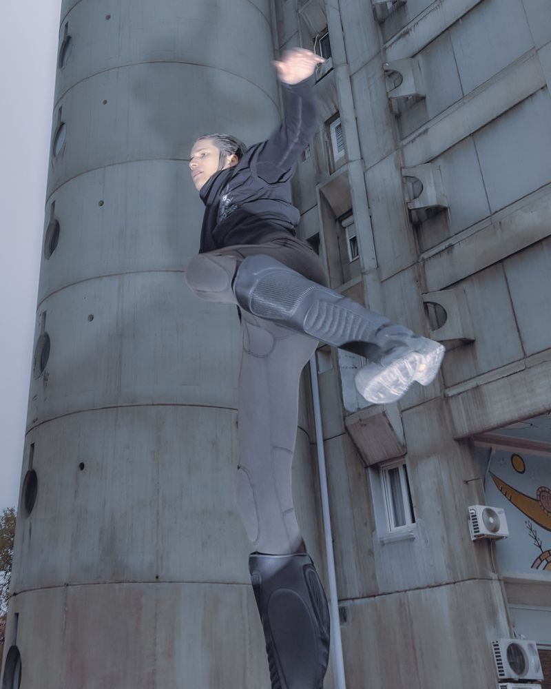 Male urban acrobat performs parkour stunt on city building, wearing activewear, with a modern architectural backdrop and overcast sky.