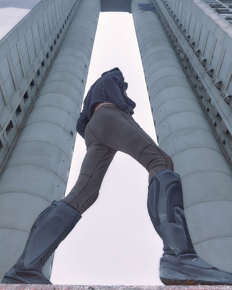 Person walking between two buildings with concrete pillars, wearing a black hoodie, gray pants, and grey and black boots.