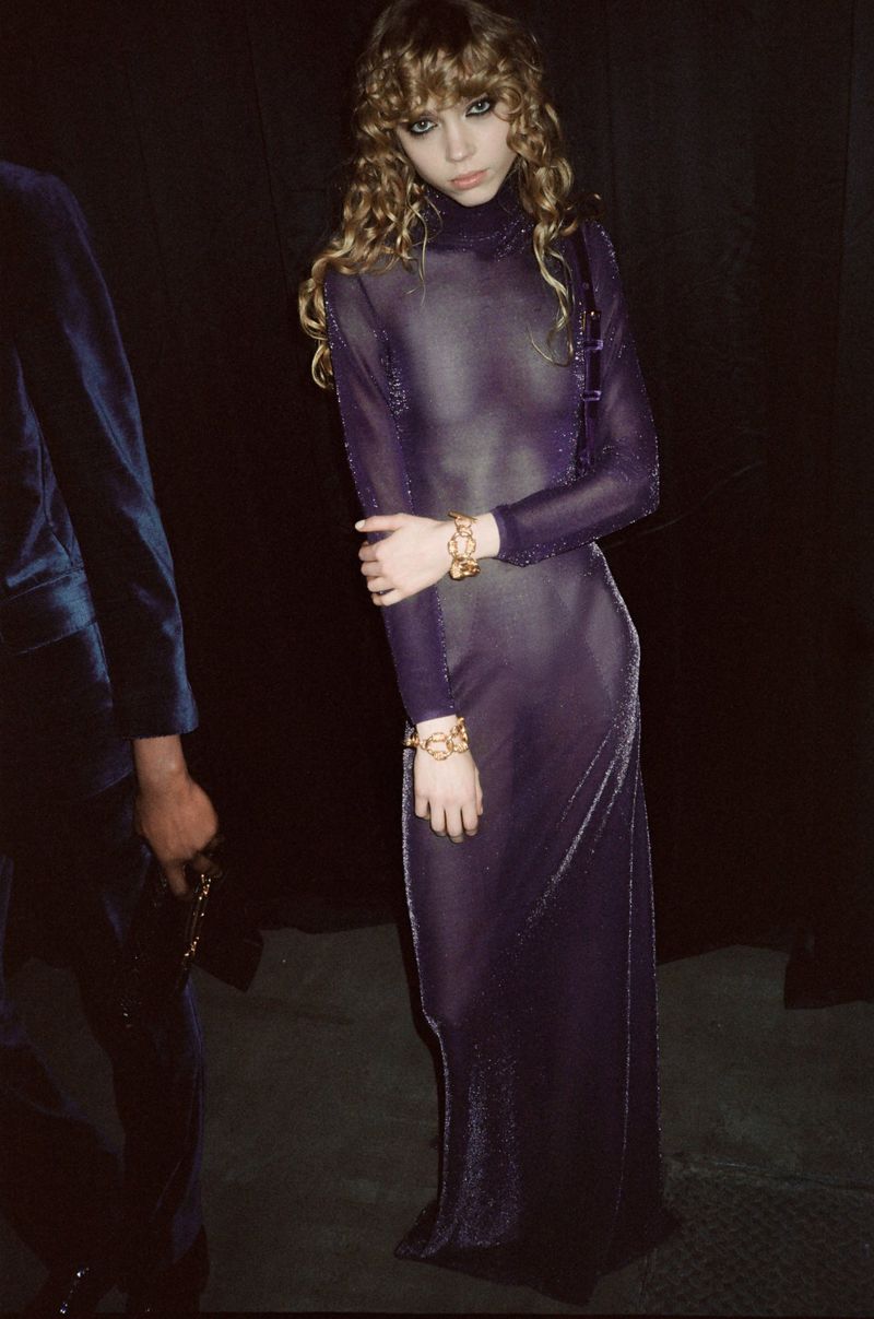 Blonde model in a sheer purple dress poses in front of a black backdrop, showcasing her slender physique and sophisticated style.