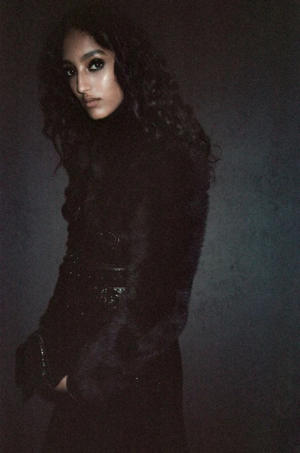 Portrait of a dark-haired woman in a dark turtleneck and patterned jacket posing in a dimly lit indoor setting with a dark background.