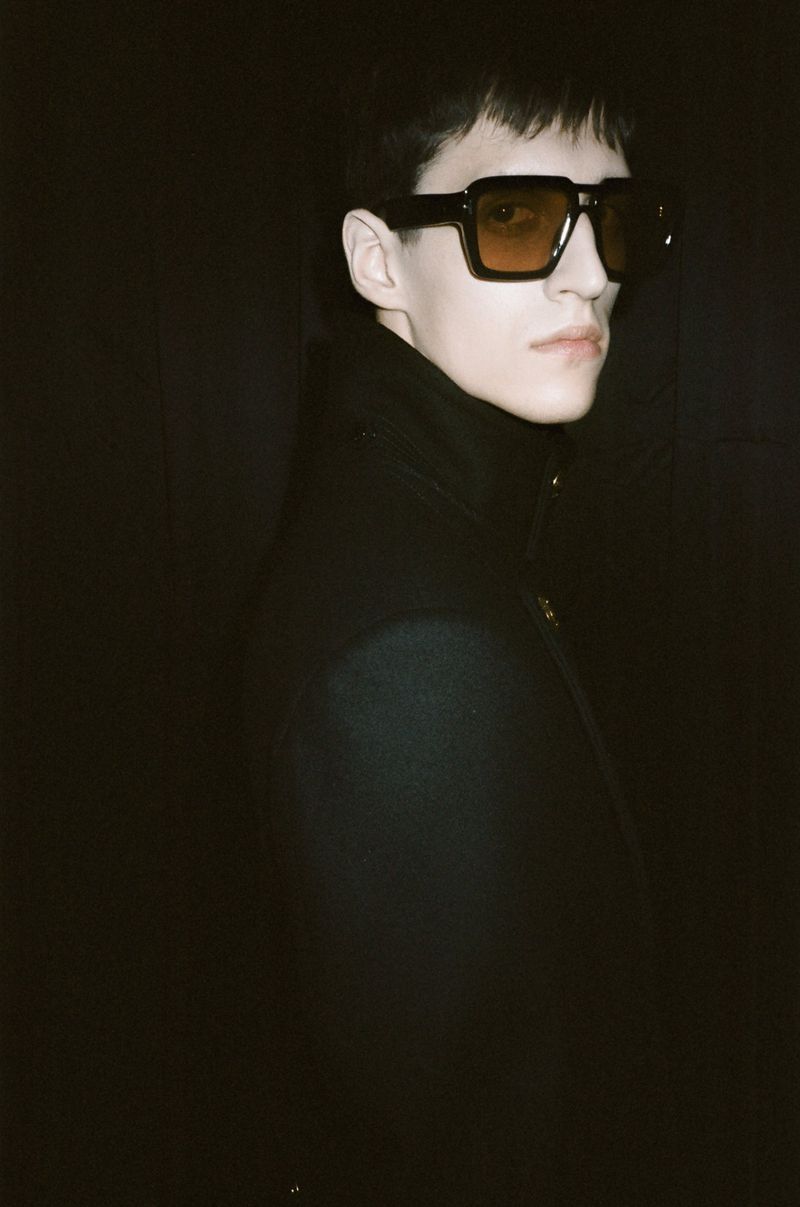 Portrait of adult male wearing sunglasses and turtleneck sweater against a dark background in a low-light studio setting.