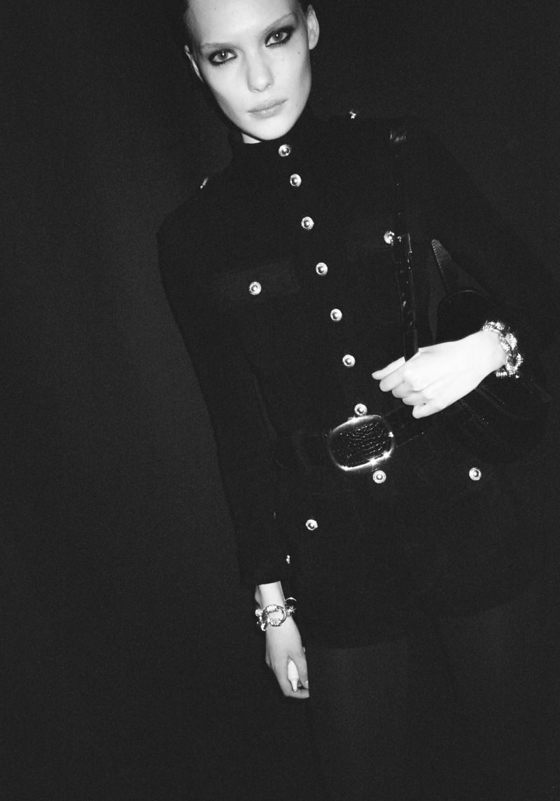 Black and white fashion portrait of a model in a fitted, military-style jacket with silver buttons and a wide belt against a dark background.