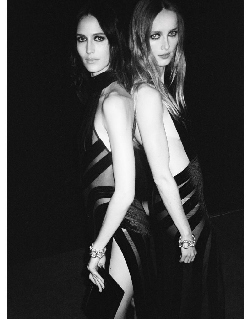 Two women in black, sheer dresses pose back-to-back against a dark background, showcasing silver bracelets and high-fashion style.