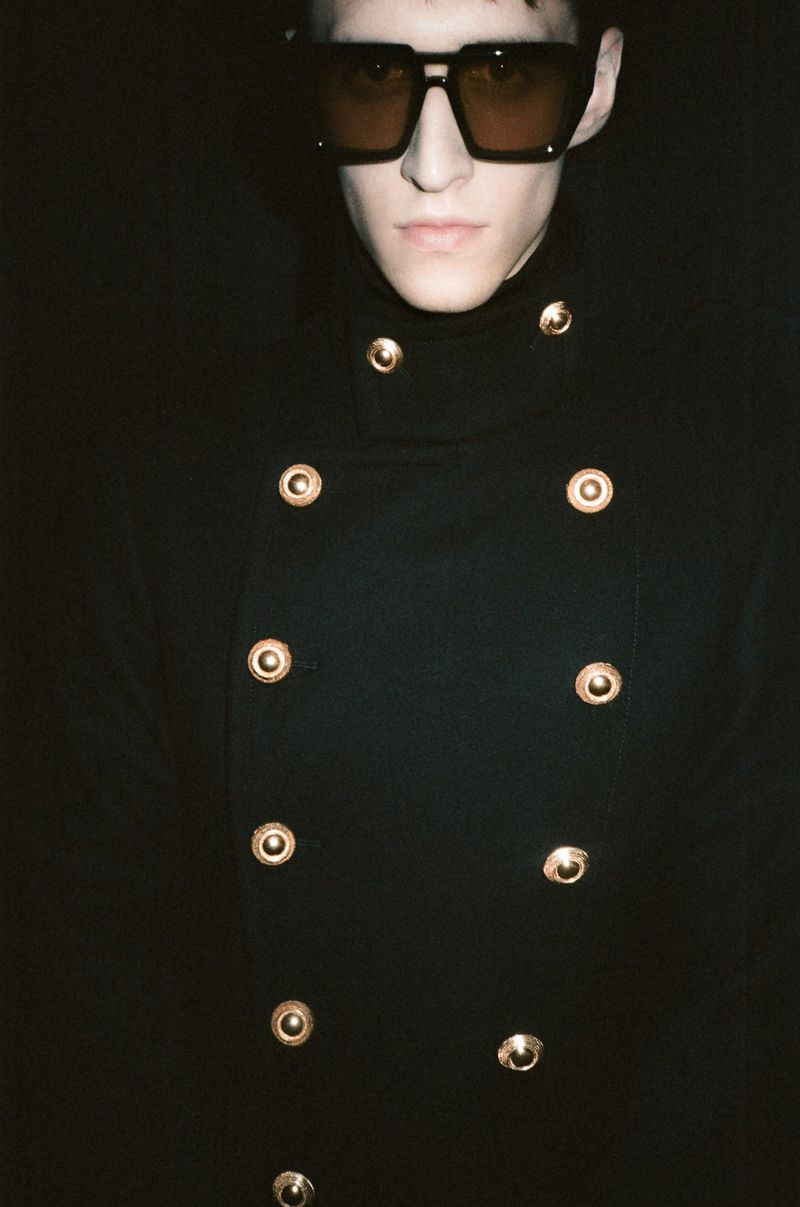 A stylish young adult in a black coat adorned with gold buttons and dark glasses stands in front of a dimly lit black backdrop.