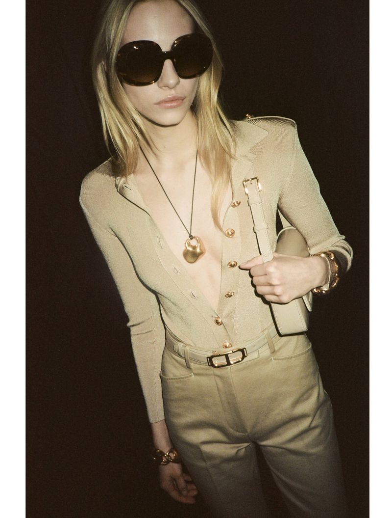 Blonde model in oversized sunglasses wearing a beige buttoned top and trousers against a dark background