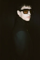 Model wearing dark oversized sunglasses and black jacket against a dark background for Tom Ford Winter 24 campaign