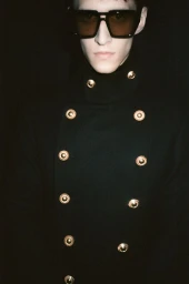 Model wearing dark oversized sunglasses and a black double-breasted coat with gold buttons against a dark background