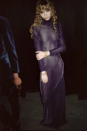 Model in a sheer purple metallic turtleneck dress with gold bracelets against a dark background.