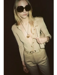 Blonde model in oversized sunglasses wearing a beige buttoned top and trousers against a dark background
