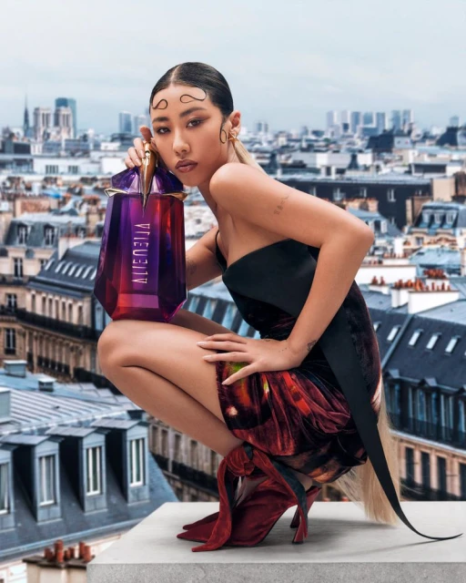 Asian model crouches in a dress, with a large purple perfume bottle, against the background of Paris cityscape and cloudy sky.