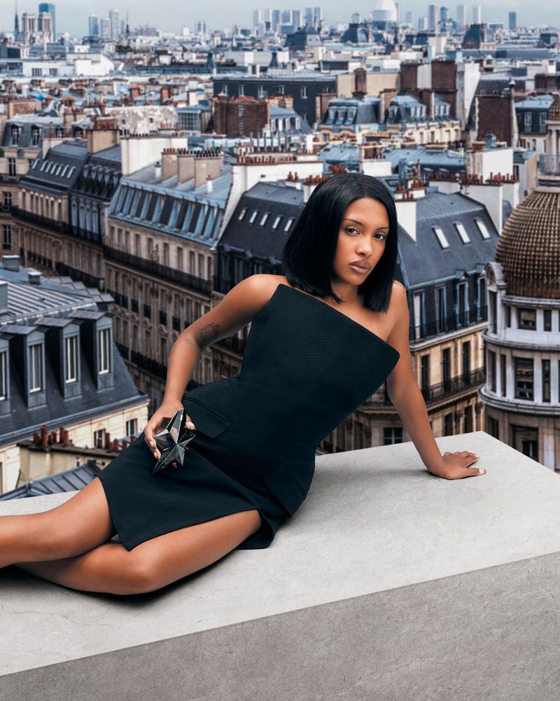 A woman in a black dress poses on a rooftop overlooking a cityscape. The model holds a star-shaped perfume bottle, styled in fashion editorial.