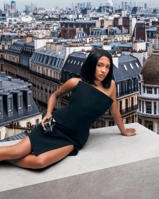 A woman in a black dress poses on a rooftop overlooking a cityscape. The model holds a star-shaped perfume bottle, styled in fashion editorial.