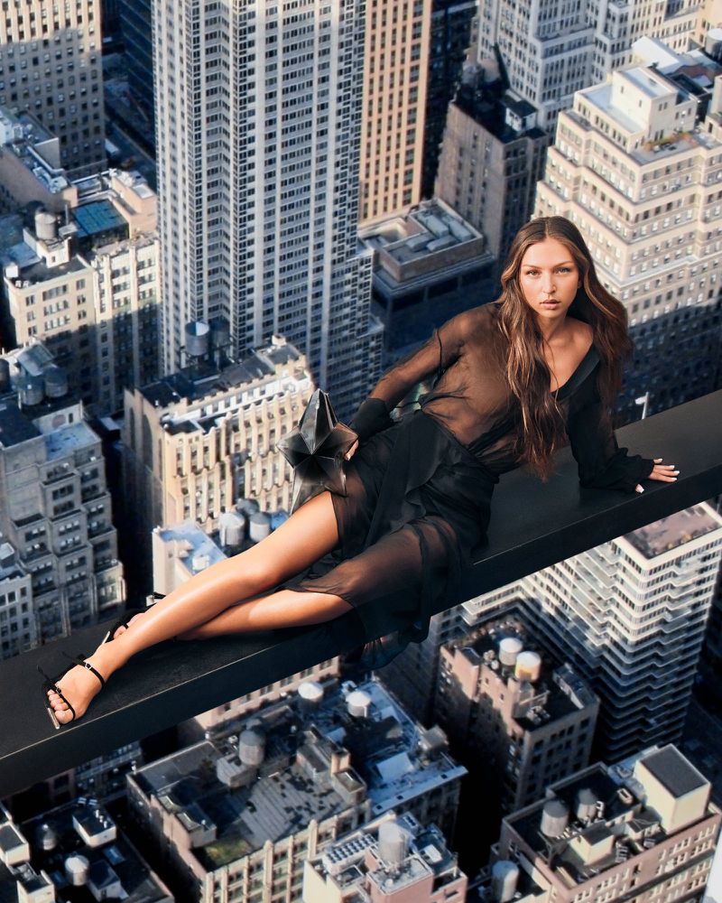 A woman in a black dress lies on a metal beam high above a city with skyscrapers in this high-angle portrait-orientation fashion shot.