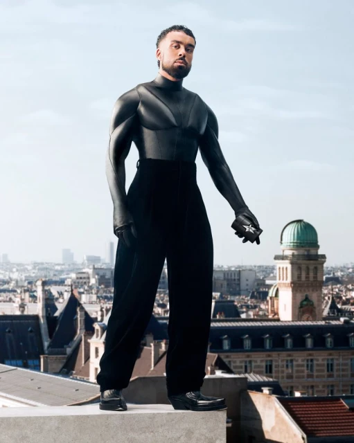 Man in modern black outfit stands on rooftop with city view in the background. The urban fashion shot captures a unique style statement.