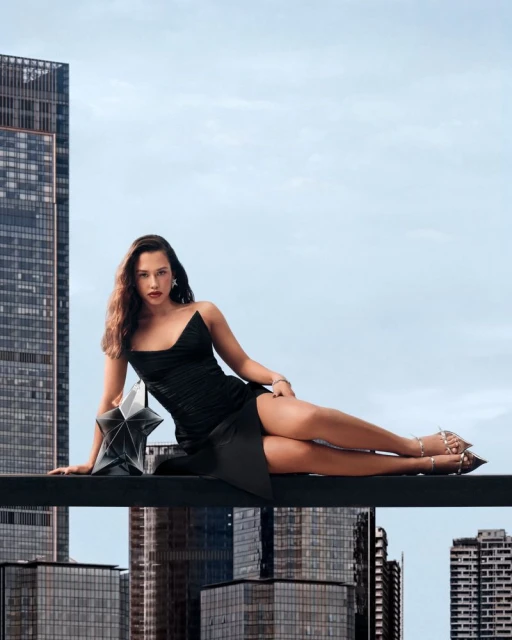 A woman in a black dress reclines on a ledge with a city skyline backdrop, accompanied by a star-shaped bag. Fashion and urban aesthetics.