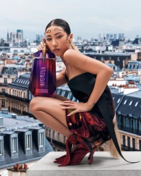 Woman in dark dress holding a purple perfume bottle on a rooftop overlooking a city skyline