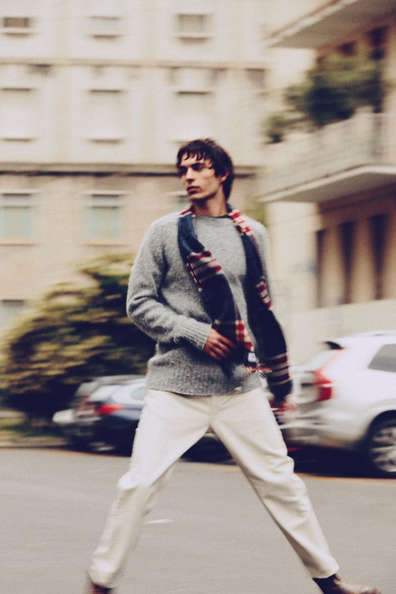 Man in sweater and white pants crosses urban street with blurred motion, buildings, and cars in background on overcast day.