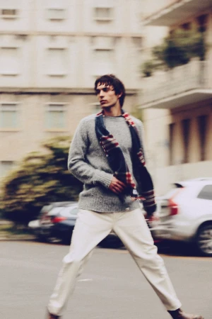 Man in sweater and white pants crosses urban street with blurred motion, buildings, and cars in background on overcast day.