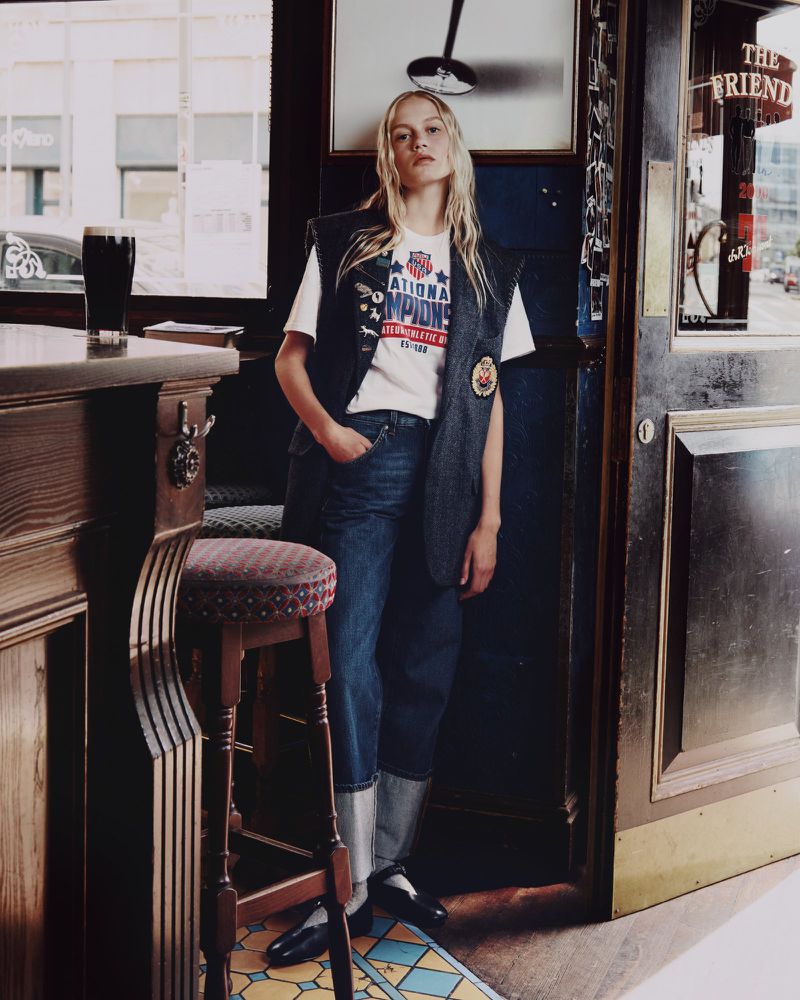 A blonde woman in a casual vintage-inspired outfit stands inside a wood-paneled bar near a stool and a dark beer on the counter.