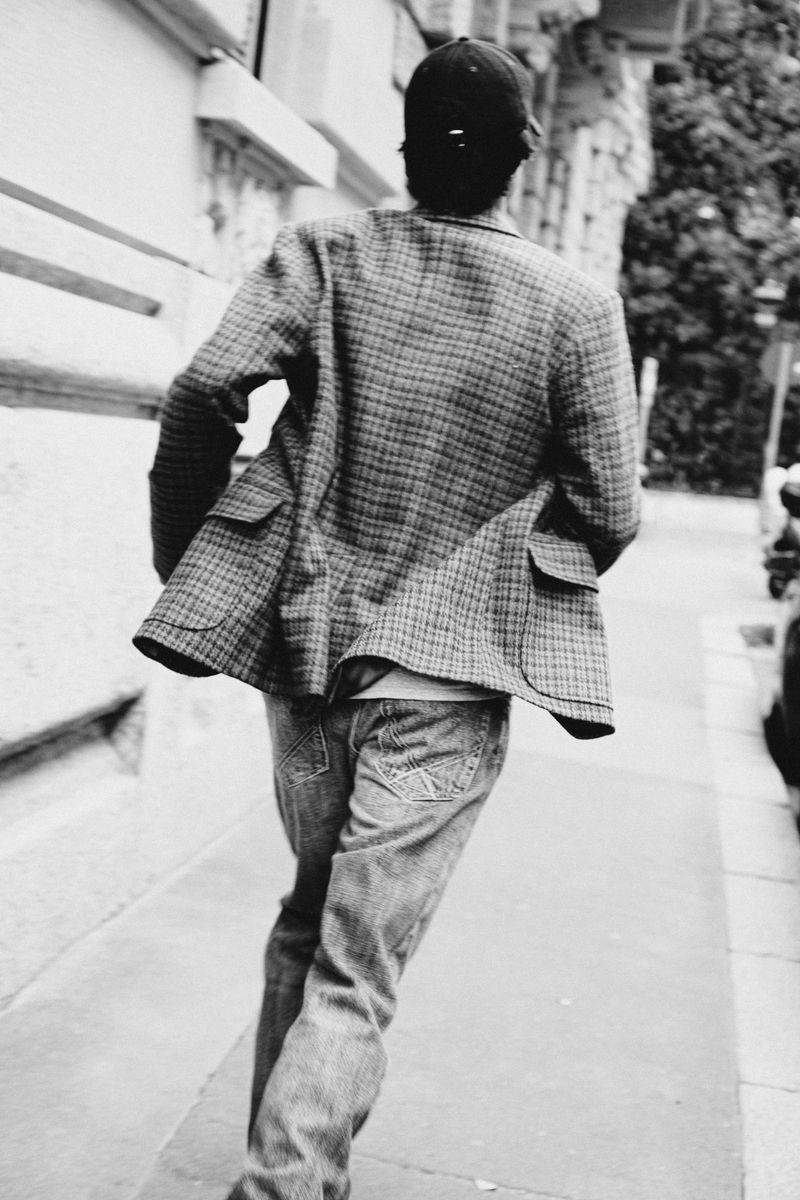 Black and white image from behind of a man in a checked jacket and jeans walking away on a city sidewalk lined with buildings and trees.